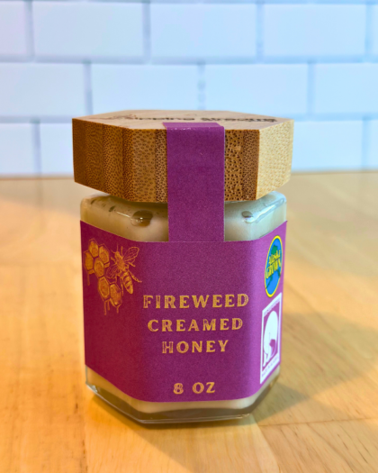 An 8 oz jar of Fireweed Creamed Honey with a hexagonal glass design and a bamboo lid. The label is deep purple with gold text and honeycomb graphics, emphasizing its Alaska-grown origin and smooth, creamy texture.