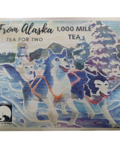 A colorful wooden postcard labeled “1,000 Mile Tea for Two”, depicting a sled dog team in a snowy Alaskan landscape. Includes two servings of tea from Sipping Streams Tea Company, inspired by the Yukon Quest race.