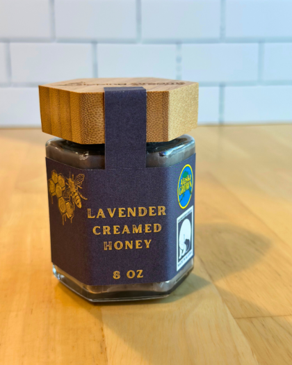 An 8 oz jar of Lavender Creamed Honey with a hexagonal glass design and a bamboo lid. The label is dark navy with gold text and honeycomb graphics, emphasizing its Alaska-grown origin and smooth, creamy consistency with a hint of lavender.
