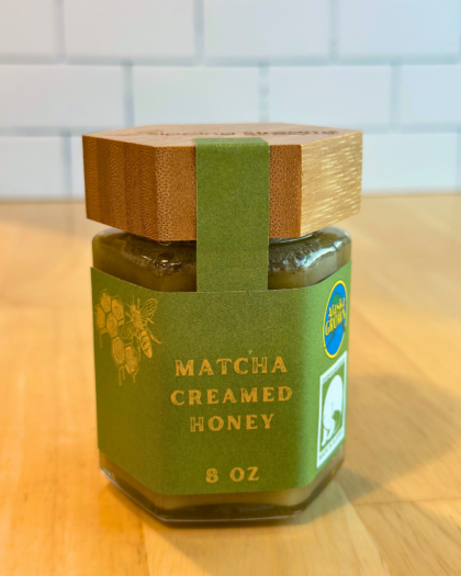 An 8 oz jar of Matcha Creamed Honey with a hexagonal glass design and a bamboo lid. The label is green with gold text and honeycomb graphics, emphasizing its Alaska-grown origin and smooth, creamy consistency blended with matcha for a rich, earthy flavor.