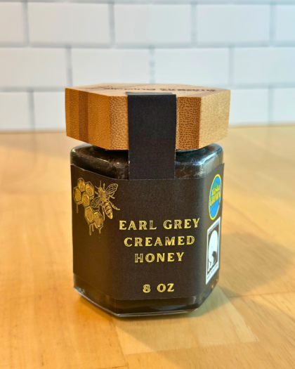 An 8 oz jar of Earl Grey Creamed Honey with a hexagonal glass design and a bamboo lid. The label is black with gold text and honeycomb graphics, emphasizing its Alaska-grown origin and smooth, creamy consistency infused with Earl Grey tea essence.
