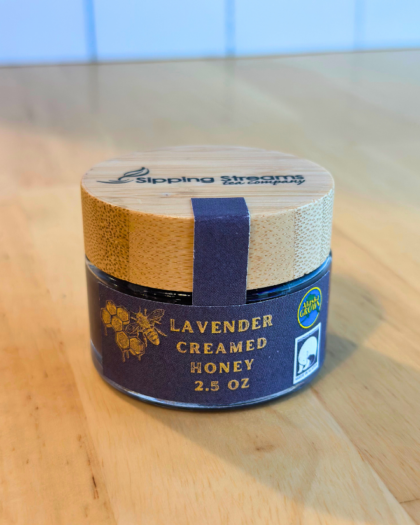 A 2.5 oz round glass jar of Lavender Creamed Honey with a bamboo lid. The dark navy label has gold text and honeycomb graphics, emphasizing its Alaska-grown origin and creamy texture with a hint of lavender.