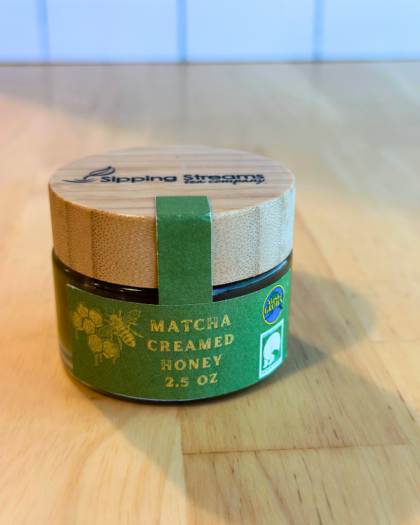 A 2.5 oz round glass jar of Matcha Creamed Honey with a bamboo lid. The green label has gold text and honeycomb graphics, emphasizing its Alaska-grown origin and creamy consistency infused with matcha.