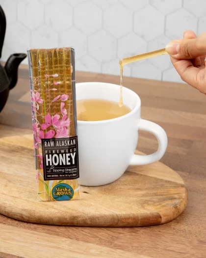 A pack of Raw Alaskan Fireweed Honey Sticks from Sipping Streams Tea Company placed on a wooden cutting board. A person is squeezing honey from a stick into a white cup of tea, with a black teapot in the background.