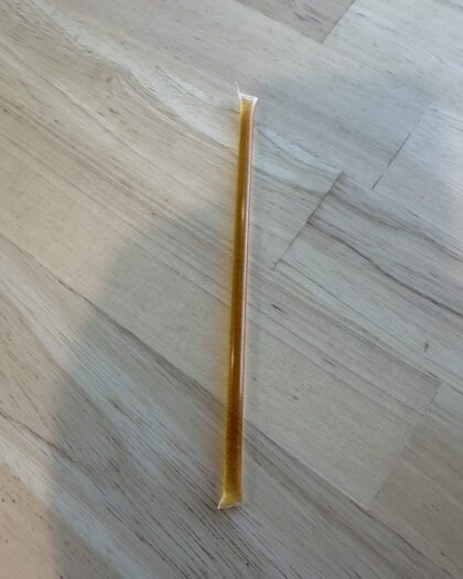 One golden honey stick filled with Alaskan Fireweed Honey, lying on a light wood surface. The honey stick contains raw, unfiltered honey known for its light floral taste and high sugar, low moisture content.