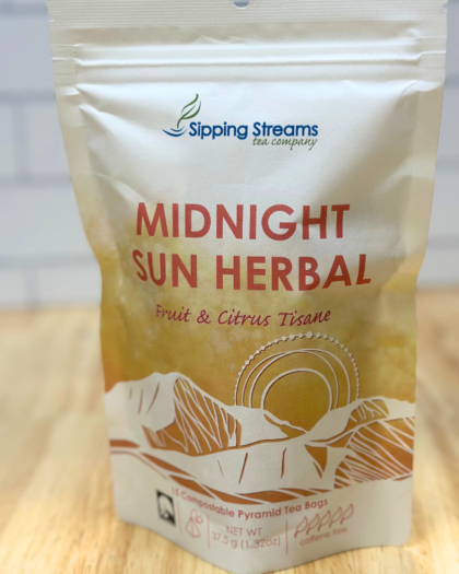 A resealable pouch of Sipping Streams Tea Company's Midnight Sun Herbal Tea. The packaging has a golden-toned mountain and sun design, indicating it is a fruit and citrus tisane with 15 compostable pyramid tea bags and is caffeine-free.