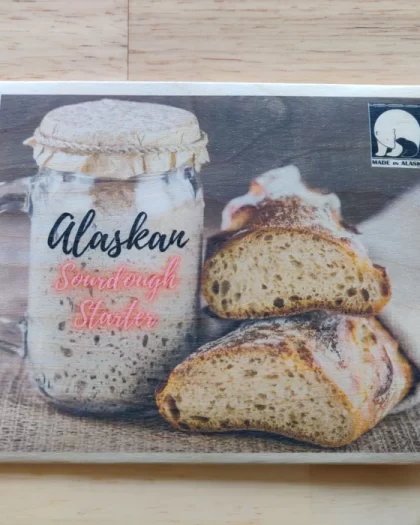 A wooden postcard showcasing an image of a jar of Alaskan sourdough starter with crusty, fresh sourdough bread. The text promotes the heritage of Alaskan sourdough, accompanied by a 'Made in Alaska' certification mark