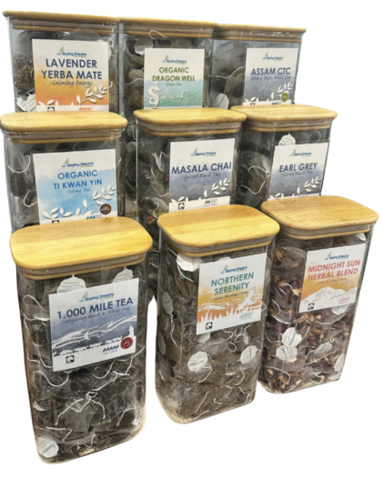 Nine clear glass jars with bamboo lids filled with individually wrapped pyramid tea sachets, arranged as a countertop tea display.